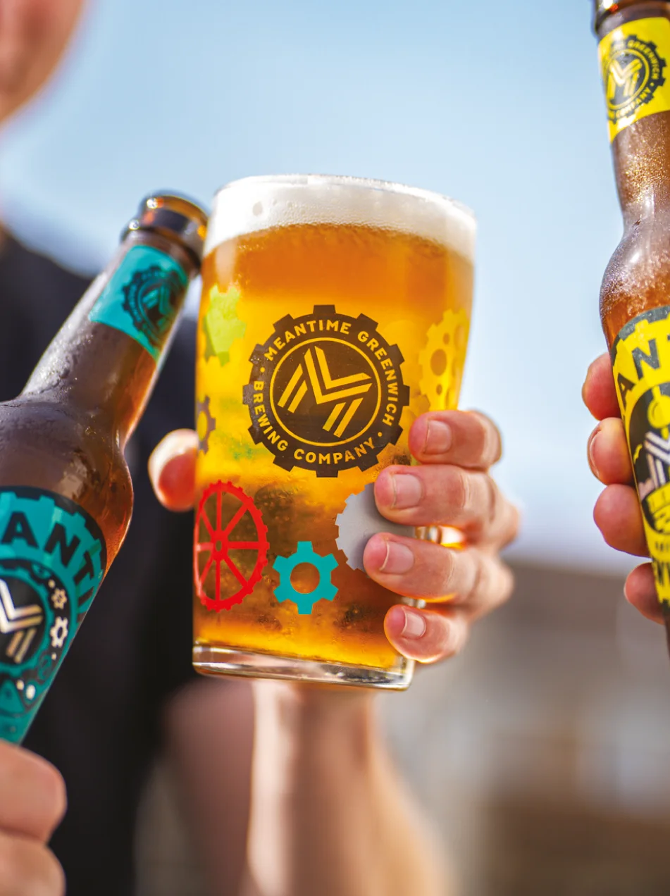 A pint of Meantime beer surrounded by two Meantime bottles clinking together against a blue summer sky